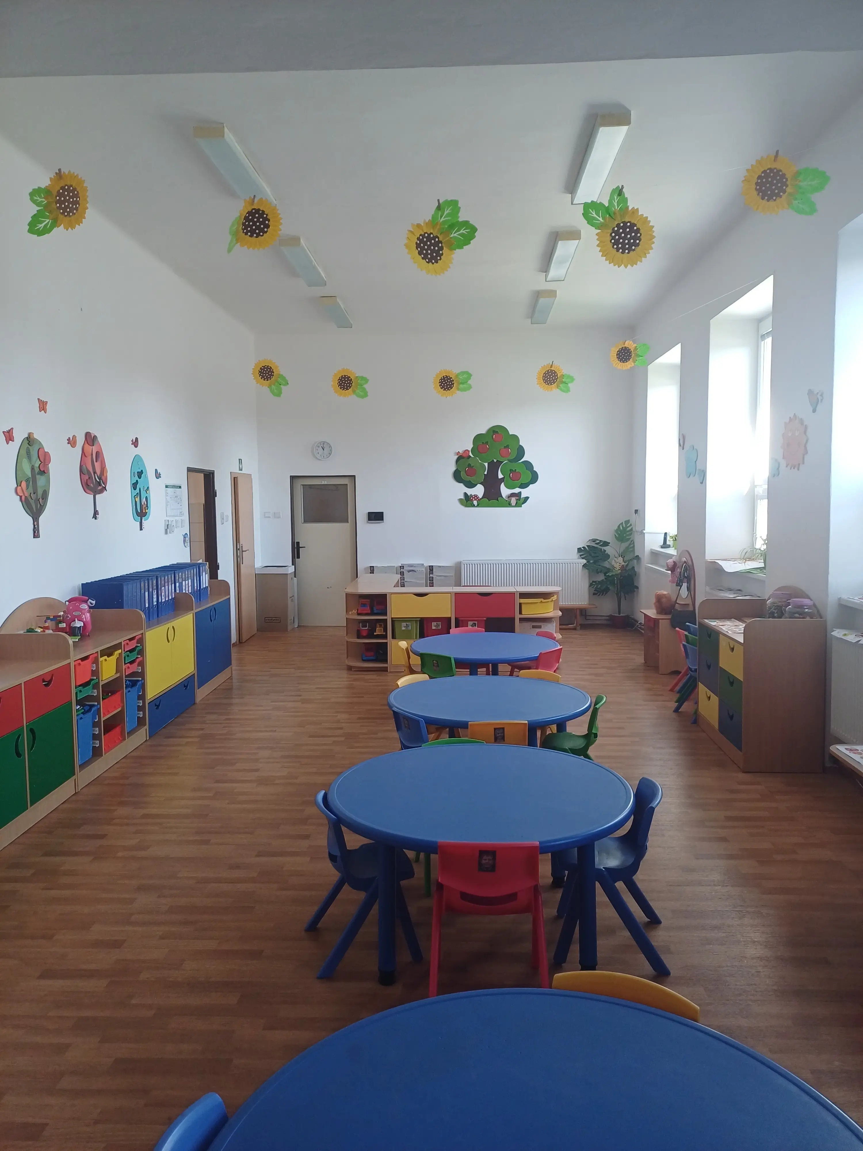 Pohled na přední část třídy s modrými stoly a židlemi a dalším barevným nábytkem vhodným pro pohyb dětí
